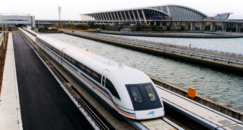 A white maglev train with a streamlined front travelling in Shanghai, China. There is a large railway station with glass walls in the background. 