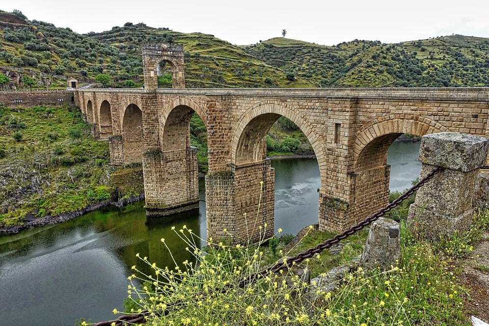 A large arch bridge with 7 arches. It is the green Spanish countryside. The river is calm as it flows under four of the middle arches