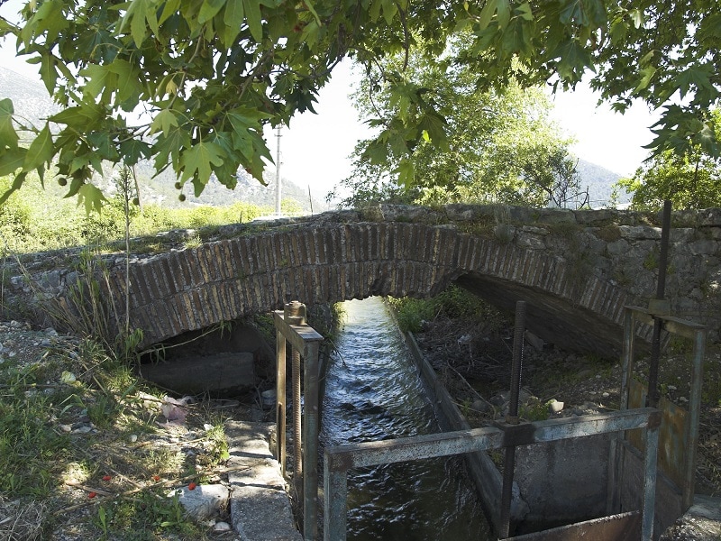 An arch bridge in a typical countryside setting. A small stream flows underneath the single arch with trees and grassy areas all around, in the far distance are two large hills.