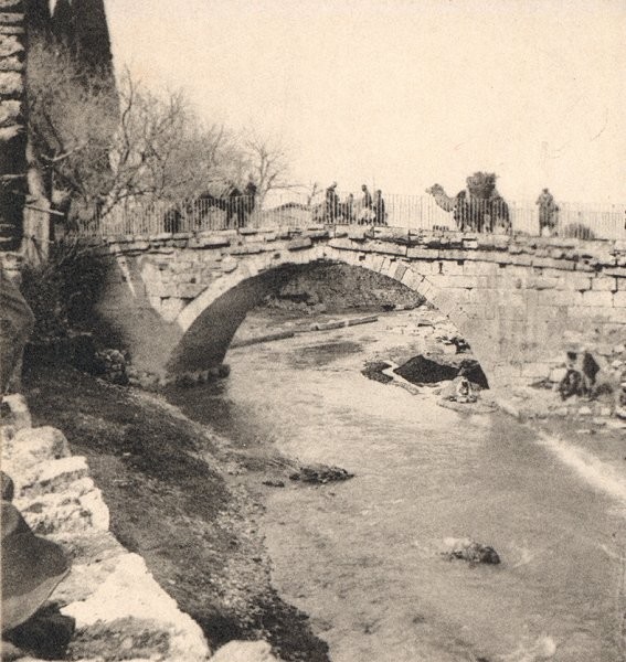 A black and white photograph of an arch bridge. There are shallow waters beneath it and camels crossing over the top under the guidance of humans 