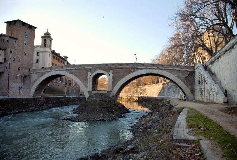 A stone bridge made up of two arches connected together. There are buildings on either side 
