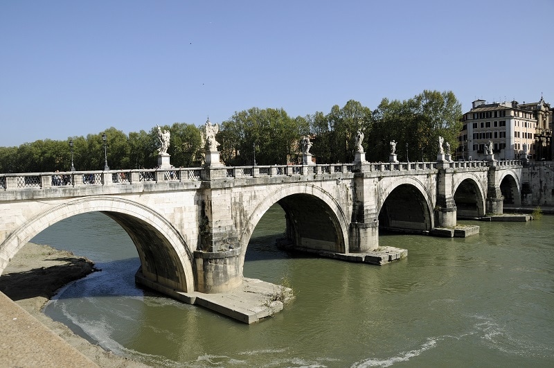 A stone arch bridge made up of 5 arches. Water flows between all of them. Lining the bridge are statues on top of the stone which connects the arches together in a way that makes them look like pillars 