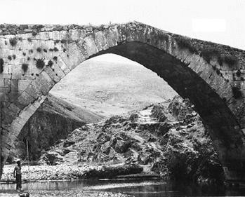 An arch bridge with a single arch. The top of the arch is pointy. A person stands underneath in the shallow waters holding a long pole. 