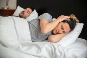 Image of a male and female in bed. Male is sleeping in the background snoring, female has ears covered and is struggling to sleep