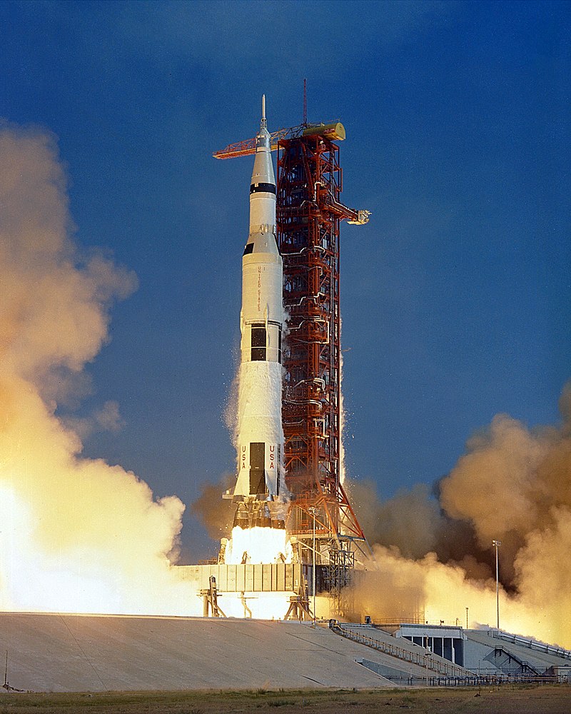 Apollo 11 stood upright on a red-framed launch pad. Flames are blasting out the three engines as it starts to go up towards space.