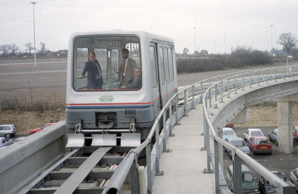 An early maglev train in Birmingham. It runs on a single track with three suited people looking towards the camera. The maglev is white with a blue and red line around the bottom. Below the raised platform the maglev is on are some parked cars.