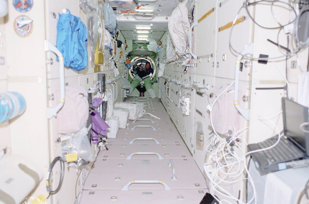 View from the interior of the Zarya module on the International Space Station. A thin corridor with handles and closed locker doors with wires in the bottom right of the picture and some white boxes on the corridor floor half way down. An astronaut can be seen without a space suit exiting a circle hatch at the bottom