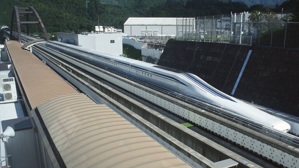 The L0 maglev train. Huge curved streamlining at the front with a blue stripe. The white maglev is running on a raised track with a station and viewing platform next to it.  