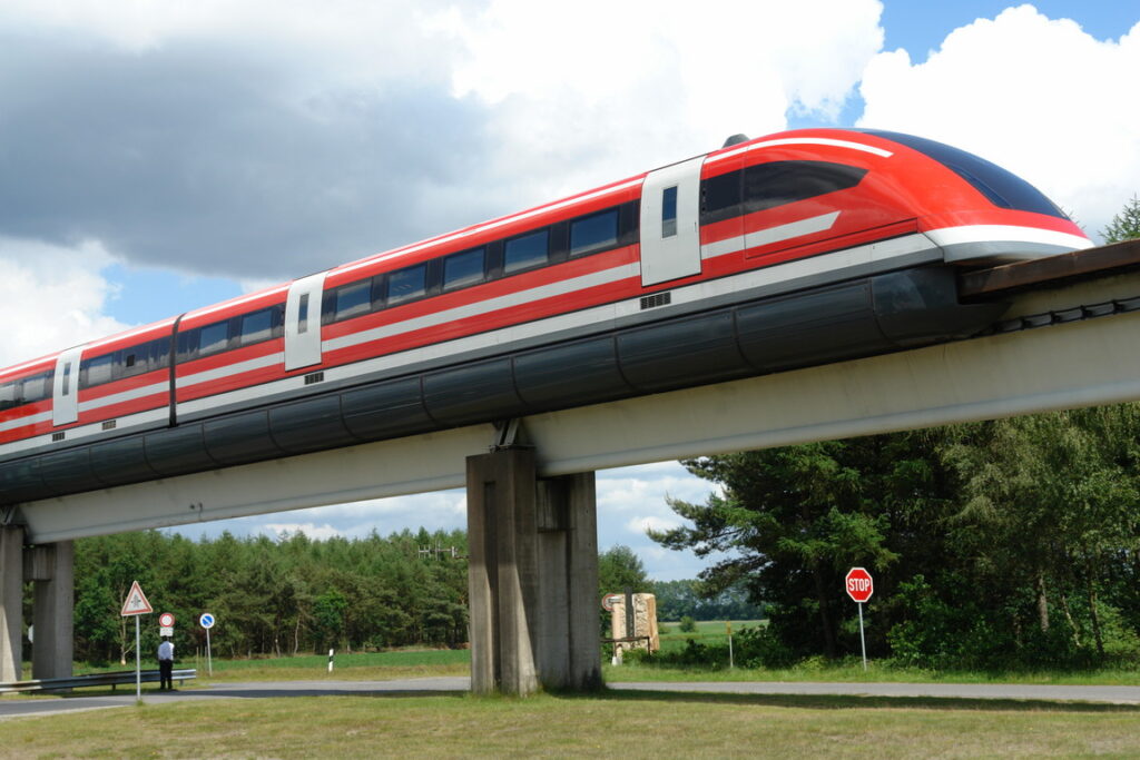 A red german maglev passing over a road and a green field. The maglev has a curved front with red paint, black windows and white doors. 