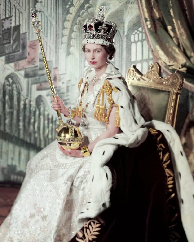Queen Elizabeth on her coronation day. She's holding the gold sovereigns orb and sceptre with a decorated cross on the end. A crown sits on her head and she wears a white dress