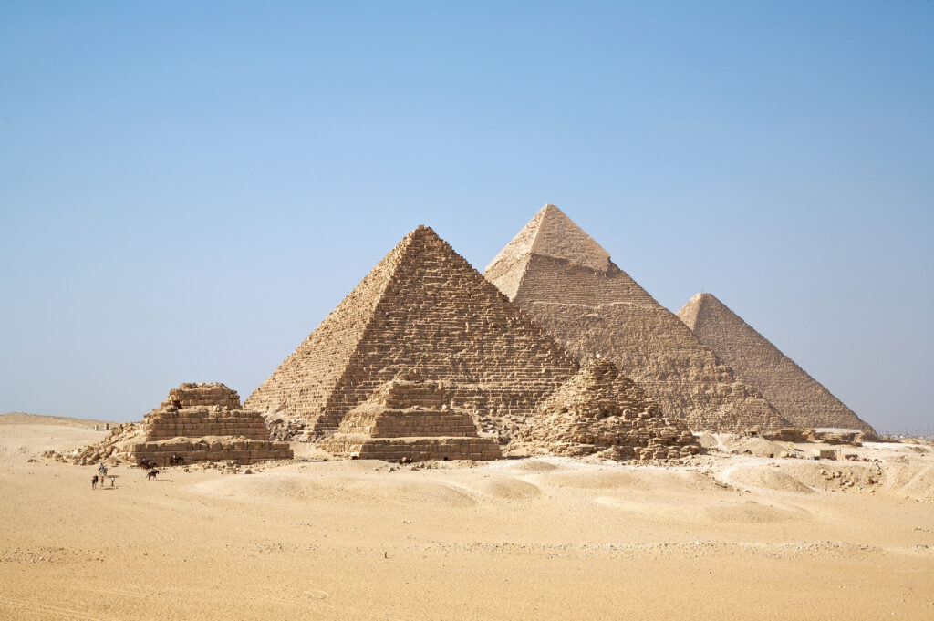 A distant shot of the three large pyramids of Giza set on sand and a bright blue sky above. One of the most notable ancient Egyptians  construction. 