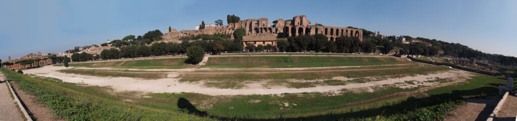 A wide shot of the remains of the Circus Maximus in Rome. It is a long with Trees line the background with ancient stone buildings where spectators would have watched sticking out. 