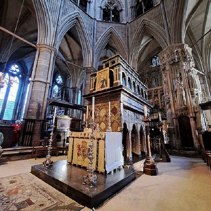 The shrine of st. Edward the confessor inside Westminster Abbey. Four silver candle sticks stand on the corners of a raised square marble platform with a table with a white cover on in front of a stone rectangle shrine. 