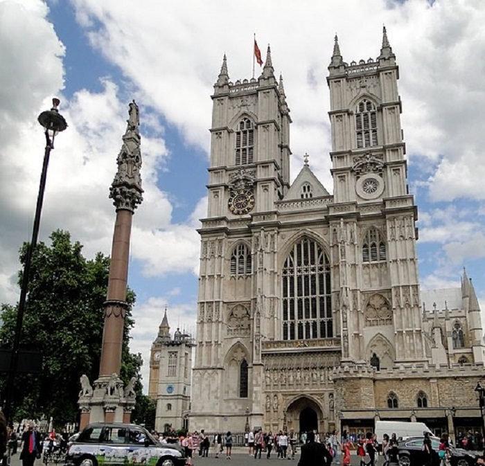 A photograph of Westminster Abbey. Tourists and locals go around their business in front of  a large arch door below a huge arch window. Two towers, one of which has a clock embedded, stretch into the cloudy sky. In the background you can see Elizabeth Tower which holds the Big Ben Bell