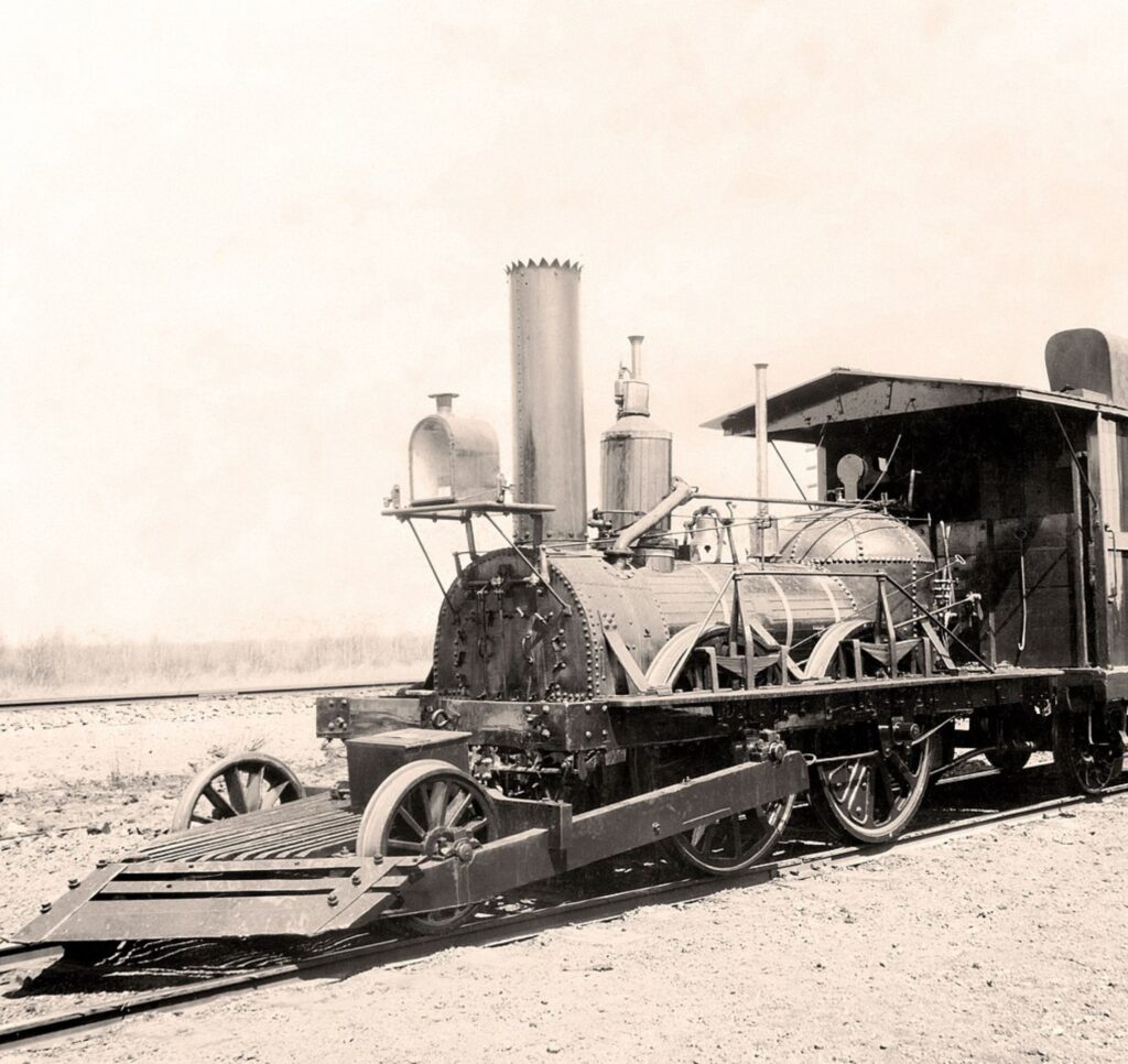 A black and white photo of the John Bull locomotive. It is on iron rails in a sandy deserted area. It has a large light in front of a tall chimney and a cab roof to protect the crew.

It has four driving wheels and two smaller wheels at the front on a connected wooden base with a cowcatcher on the front. 