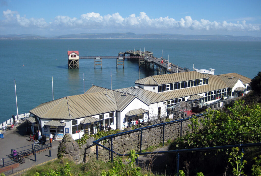 Overlooking the Mumbles pier. A long white building which used to be a railway station with glass windows spaced across. A wooden pier stretching out into the ocean behind. 