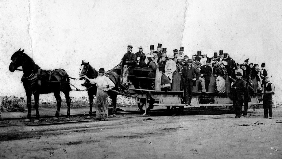 Image of two horses pulling a wooden trailer on rails. Lots of people dressed in Victorian clothing are sat on the trailer with one person guiding the horses.