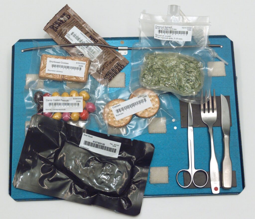 An astronauts meal. There is a pair of metal scissors, fork and knife stuck on a magnet in the bottom right, and food (including beef steak and spinach) in freeze-dried bags. 